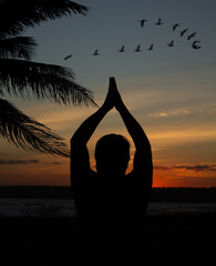 Silhouette of adult male praying at sunset with birds flying in the sky.