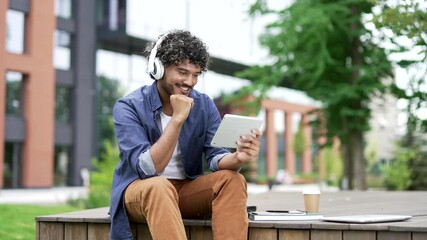 Male student watches video streaming on digital tablet while sitting on bench in campus near university building. Smiling handsome man with headphones listens to a podcast, broadcast, browses the web - Powered by Adobe