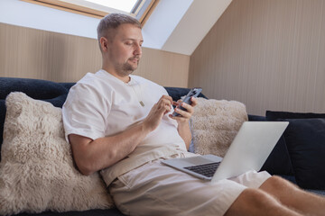 Young man sitting on sofa with laptop and smartphone, working remotely or managing online tasks. Concept of freelancing, digital lifestyle, mobile technology, e-banking, online shopping, communication