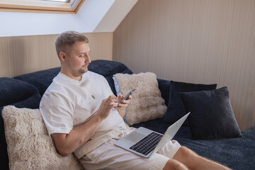 Young man sitting on sofa with laptop and smartphone, working remotely or managing online tasks. Concept of freelancing, digital lifestyle, mobile technology, e-banking, online shopping, communication