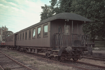 Obraz premium Vintage wooden railway carriage standing on old tracks near industrial buildings and green trees under cloudy sky