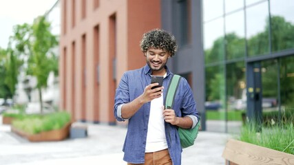 Smiling student with a backpack uses a mobile phone while walking in the campus space near university building. Handsome man chats online with friend, browses social media, writes or reads messages