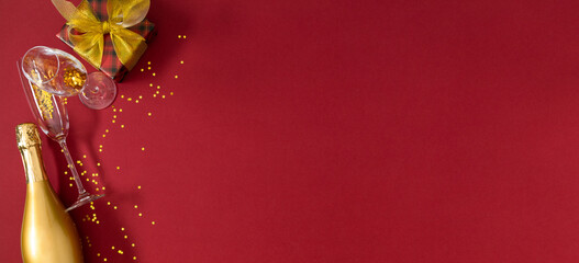 Golden bottle of champagne, glasses and a golden gift, glitter and confetti on a dark red...