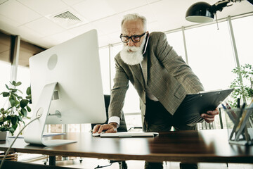 Elderly businessman analyzing data at office, multitasking with computer and documents, demonstrating confidence and professionalism
