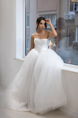 Naklejka premium Elegant bride in crystal embellished ball gown, sitting gracefully near window indoors