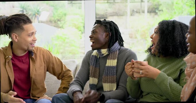 Diverse friends laughing after scarf wearing woman handing ceramic mug on sofa to share drink