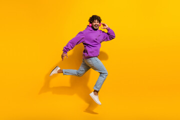 Energetic young man in purple hoodie and jeans jumping with joy against bright yellow background, showcasing vibrant energy
