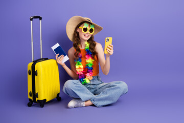 Cheerful girl traveling with her summer suitcase, passport, and smartphone against a vibrant purple background