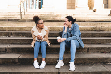 Two young women sit on stone steps, talking and drinking coffee together on a bright afternoon