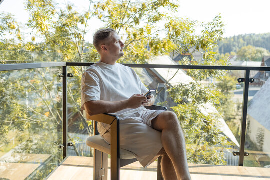 Casual man in white clothes sitting on balcony with coffee cup and smartphone, enjoying peaceful outdoor view of trees and houses. Concept of relaxation, modern lifestyle, and digital connection.