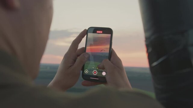 Rear view of lady recording scenic sunset and distant paraglider using smartphone while riding in hot air balloon over countryside, glowing sky and open landscape