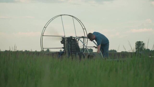 young man in blue shirt leans over paramotor on grassy field at dusk starting engine causing fan blades to spin rapidly within metal cage against open farm sky and ready for flight