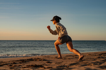 Athletic woman training on sandy beach at sunset, performing deep lunge exercise in gray shorts, sweatshirt, and cap, with focused posture and ocean horizon in the background