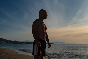 Strong bald man standing shirtless on sandy beach at sunset, holding resistance bands in hand, looking thoughtfully at the sea horizon with mountains and dramatic sky in background
