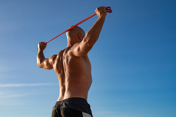 Strong athletic man exercising with resistance band outdoors, back view of muscular shirtless male...