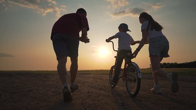 Boy learns to ride with help from mother and father holding his bicycle. Family supports training on open path at sunset. Sunset silhouettes child, bike, and parents in active evening family learning.