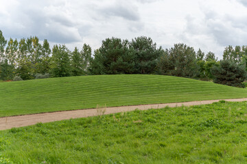 Obraz premium A smooth green hill rises under cloudy skies. Neatly trimmed grass contrasts with the surrounding trees.