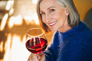 Smiling mature woman enjoying a glass of red wine indoors, wearing a cozy sweater during a relaxing day at home.