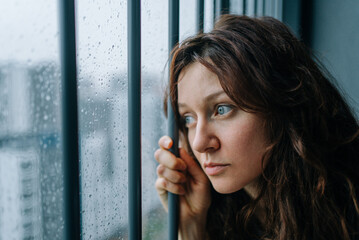 Young woman staring through a rain-streaked window, feeling lonely and melancholic, contemplating...