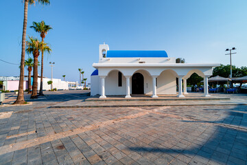 White Greek-style church building on street with palm-lined square in port Ayia Napa, Cyprus. Sightseeing in Mediterranean city on sunny day.