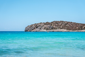 Obraz premium Crystal clear turquoise waters meet a rocky, shrub covered hill extending into the sea on Crete's coastline, with a vibrant blue sky above.