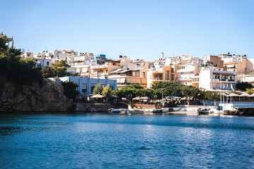 A Mediterranean town Agios Nikolaos on Crete, Greece, with white and pastel buildings by calm turquoise water. Boats dock near the shore, with greenery on a rocky outcrop.