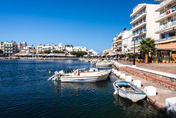Calm turquoise water with small boats, a promenade with cafes and palm trees, and pastel colored buildings under a bright blue sky in Crete, Greece.
