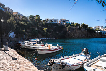 A tranquil harbor on Crete, Greece, with fishing boats moored by a stone paved waterfront. Turquoise water reflects sunlight, with cliffs and buildings in view. © Aerial Film Studio