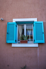 A turquoise window with open shutters on a terracotta wall, featuring lace curtains, a potted plant, and a security camera, in Crete's Mediterranean style.