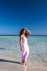 A woman in a light pink dress stands in shallow turquoise water on a sunny day. The serene beach on Crete features white sand and calm waves.