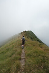 Atmospheric scene of a person walking along a narrow path on a mountain peak, surrounded by dense fog and mist. The dramatic landscape conveys solitude, adventure, and the mysterious beauty