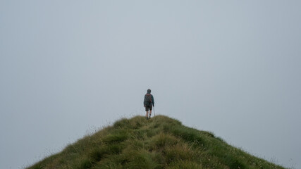 Atmospheric scene of a person walking along a narrow path on a mountain peak, surrounded by dense fog and mist. The dramatic landscape conveys solitude, adventure, and the mysterious beauty