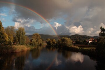 Vibrant rainbow arcs across a dramatic sky over a tranquil river and lush landscape.