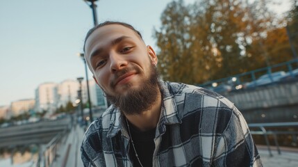 A man with a beard and plaid shirt stands outdoors, surrounded by buildings and trees. The man leans forward slightly, near the bridge as cars pass by buildings.
