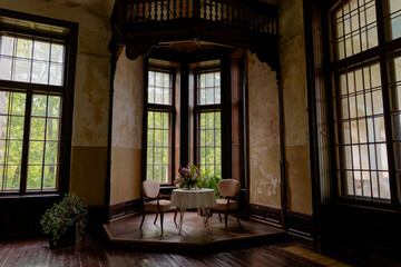 An old, empty room with tall windows and a small table set for two, decorated with a flower bouquet.