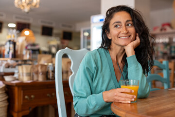 Happy woman with long black curly hair wearing a turquoise sweater, sitting at a cozy caf&eacute; table and smiling while holding a glass of fresh orange juice with a black straw