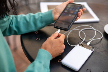 Close up of woman connecting smartphone to portable power bank with charging cable on table indoors, modern technology and battery life solution concept.