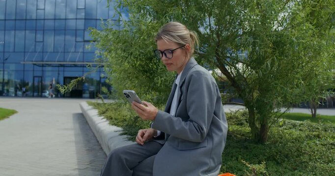 A businesswoman uses a mobile phone sitting on a parapet on a city street. A businesswoman uses a mobile phone near an office building.