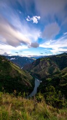 High-angle view of a winding river valley. Lush green hills and mountains encircle a curving river snaking through the valley.  A mix of light and dark clouds graces a vibrant blue sky