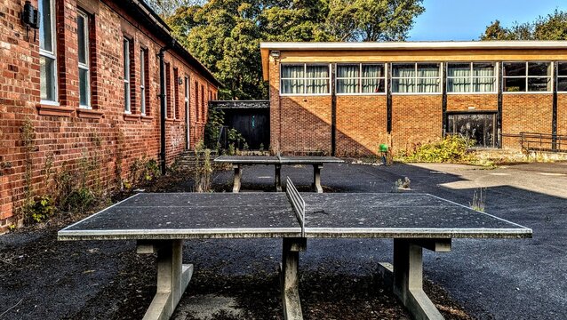 Derelict outdoor ping-pong sports equipment, concrete table tennis, deserted playground. Post-apocalyptic scene, failing crumbling education system school-yard. Neglected empty buildings, courtyard.