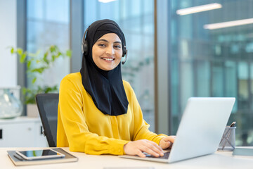 Muslim woman wearing a hijab and headset, sitting at an office desk, smiling at the camera while typing on a laptop, providing professional customer service support