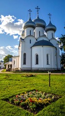 Orthodox church with domes,  a sunny day.  Exterior view of a  white church  with  multiple  rounded domes.  A grassy  area surrounds the building,  with small flowerbeds.  Bright sky