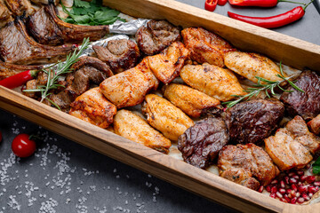 Grilled meat platter with roasted chicken wings and beef. Wooden serving platter with grilled chicken wings, beef, pork, herbs, vegetables and spices on a gray surface.
