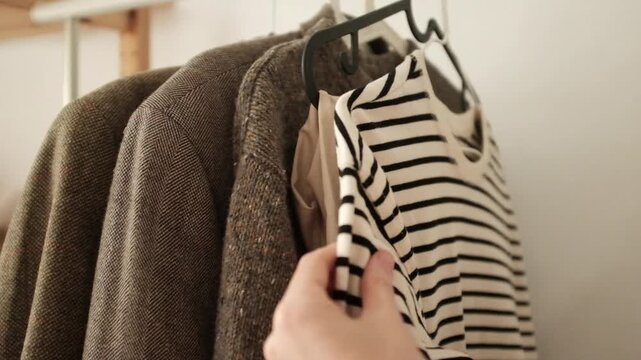 Young adult woman selects or organizes clothes on a clothing rack. Preparing for a cold season. Choosing blazers and sweaters. Second hand thrift sale.  Concept of fast fashion industry waste issue. 