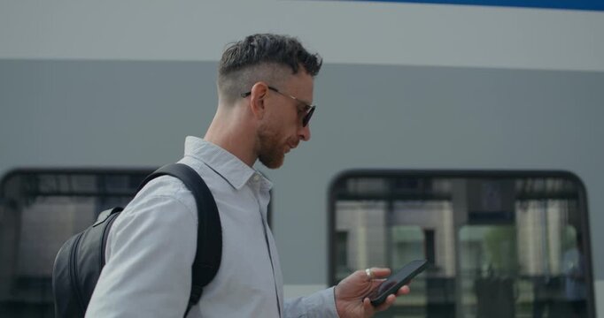 A man with backpack sends a text message on his mobile phone and drinks coffee from a disposable cup while walking along the train at the platform.