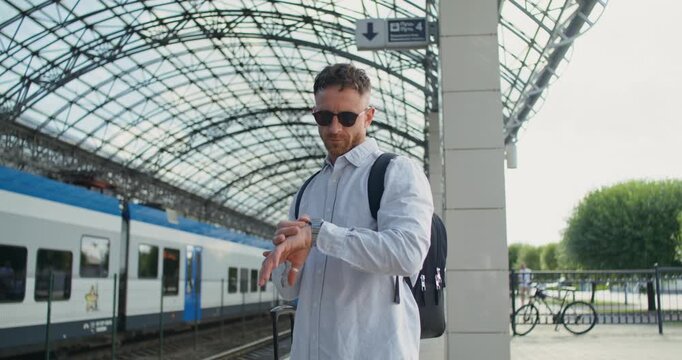 A man looks at his wristwatch standing on the station platform with a suitcase while waiting for a train.