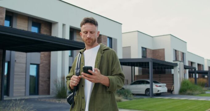 A man with a backpack watches the time on his mobile phone walking down the street past townhouses.