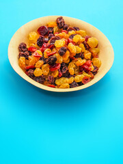 A bowl of dried fruit with yellow and red raisins, cranberries. The bowl is on a blue table surface. Natural high sugar and glucose content. Popular snack.