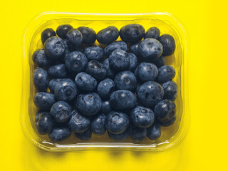 A plastic tray of blueberries sits on a bold yellow color background. The tray is clear and filled with fresh organic fruits ready for consumption. Healthy diet concept. Natural product.