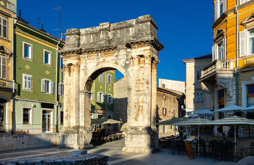 Obraz premium Ancient Arch of Sergii in Pula, Croatia, lighted by bright summer sunlight under clear blue sky, blending with narrow stone streets, colorful houses, and outdoor cafes in lively Mediterranean setting